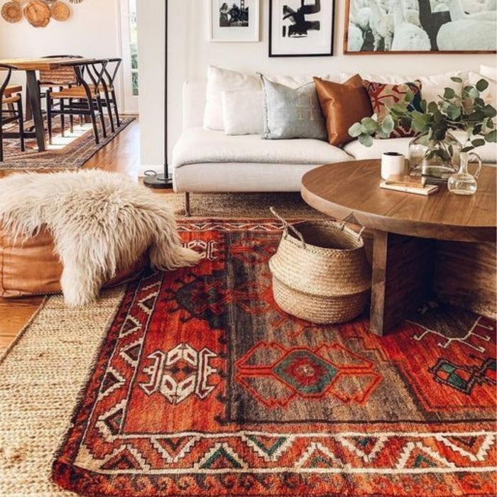 A warm living room featuring a vibrant vintage red kilim rug with intricate geometric designs, a white sofa with neutral-toned pillows, a round wooden coffee table, and natural accents like a woven basket and a faux fur pouf.