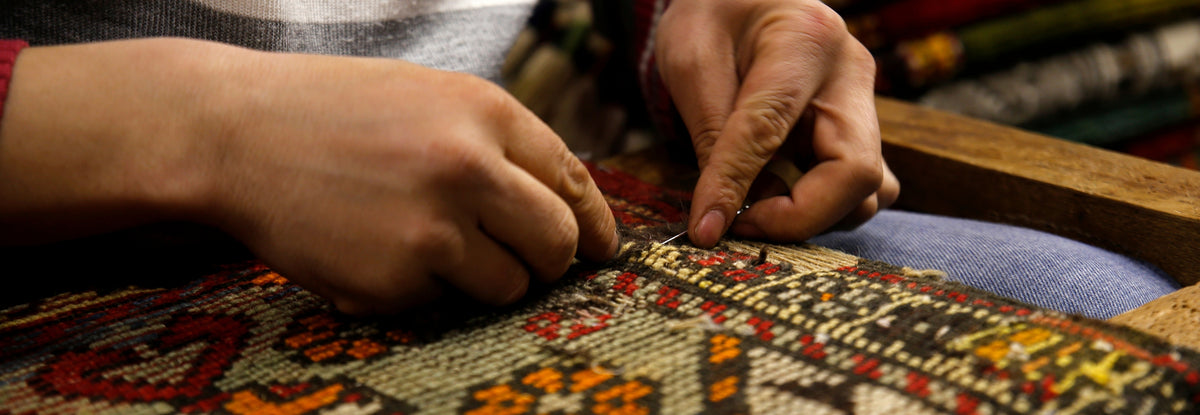 A man repairing a vintage rug, carefully restoring its intricate patterns and fabric. He is focused on his work, seated on the floor with tools in hand, as he carefully mends the delicate threads and motifs of the aged carpet.
