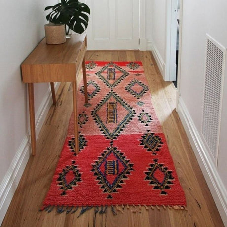 A vintage kilim runner rug with black diamond patterns on a rich red base lies elegantly in the hallway. On the left side, a wooden table rests against the wall, topped with a decorative potted plant. The hallway walls are painted crisp white, creating a clean and minimalist contrast to the rug’s bold design.