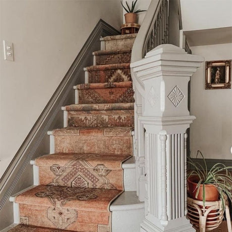 A vintage runner rug with traditional motifs and pastel colors is laid out on a white staircase. On the right side of the stairs, there is a white planter, complementing the soft hues of the rug and enhancing the serene atmosphere of the space.