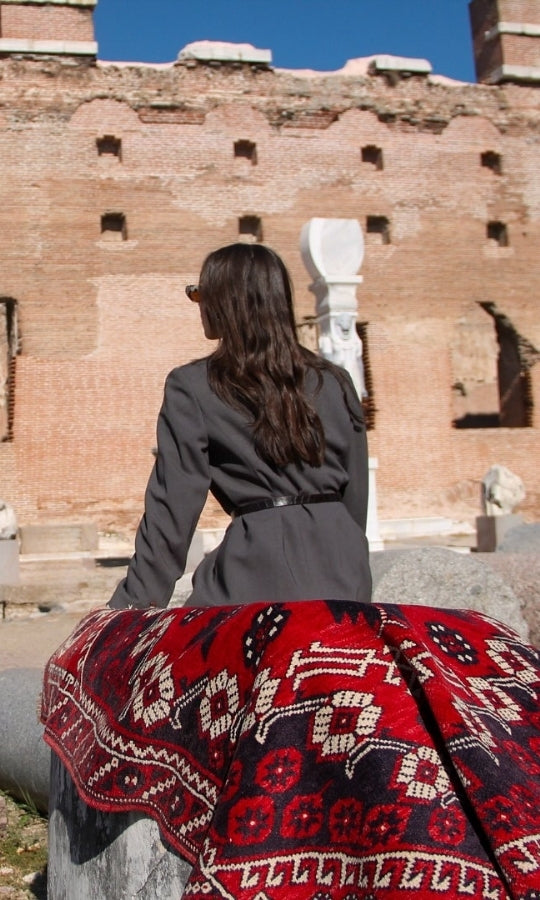 A woman in gray attire and glasses sits on a vintage rug with traditional red motifs in an ancient ruin, gazing into the distance.