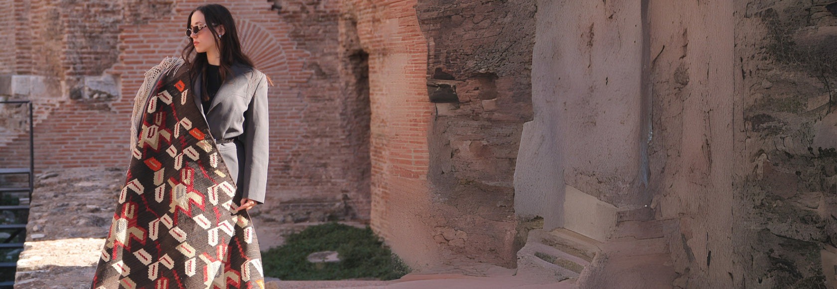 A woman in gray attire, wearing glasses, holds a traditional beige and brown vintage rug with intricate motifs. She poses amidst the ruins of ancient architecture, the worn stone walls and historical elements creating a striking contrast with her modern, minimalist look.